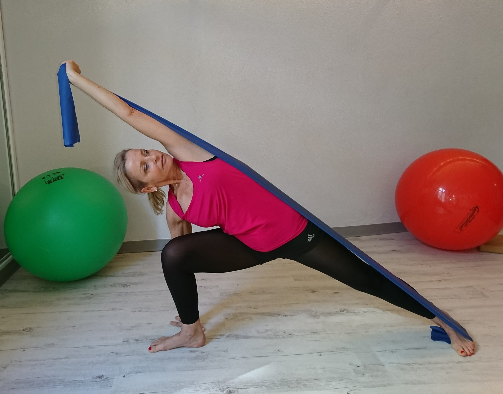 Une femme en haut rose et legging noir effectue une fente latérale en s'étirant avec une bande élastique bleue, entre deux gros ballons de gymnastique vert et rouge.