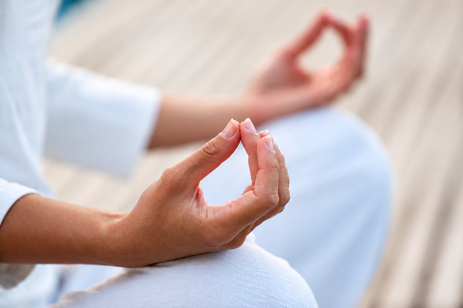 Gros plan sur les mains d'une personne assise en méditation, le pouce et l'index joints, vêtue de blanc.