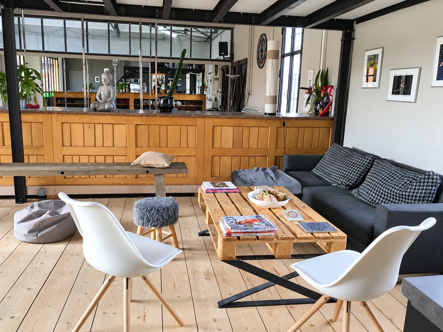 Un espace salon de style loft avec une table basse en palettes, des chaises blanches, un canapé gris foncé et un comptoir en bois à l'arrière-plan.